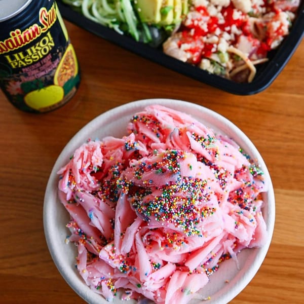 Pink shave ice with colorful sprinkles, a poke bowl, and a Hawaiian Sun drink on a wooden table.
